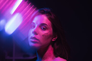 Cyberpunk photo of attractive woman in colored light on dark background with glitter on face posing...