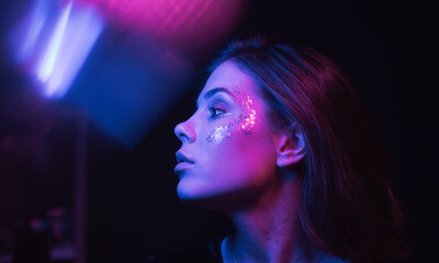 Neon cinematic portrait of a brunette woman with bright makeup in a dark room with purple and blue light. Trendy portrait of a woman in glitter with makeup.