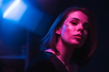 Trendy night portrait of a beautiful woman with beautiful makeup in the style of euphoria, posing in a dark room in neon light, looking at the camera.