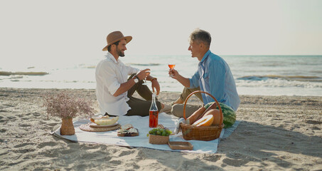 European happy gay couple drinking wine and enjoying romantic picnic at beach. Homosexual...