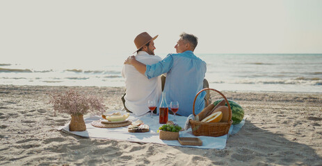 Homosexual lgbt couple, gay men having picnic at the beach, sitting on blanket and embracing,...