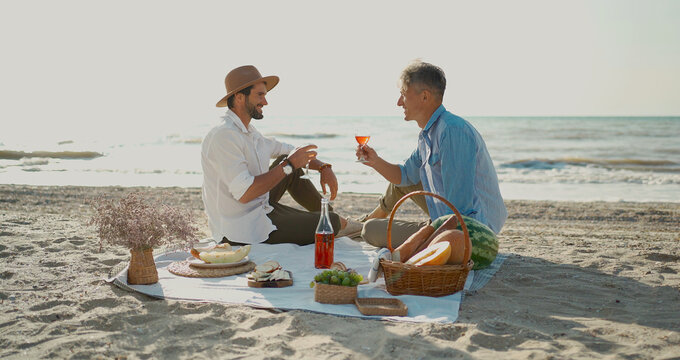 European happy gay couple drinking wine and enjoying romantic picnic at beach. Homosexual relationships and alternative love lifestyle concept