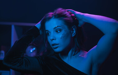 Close portrait of woman in makeup with glitter in blue light in dark room, looking aside. A woman...