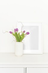  bouquet of tulips in a white vase on a white chest of drawers and empty frame