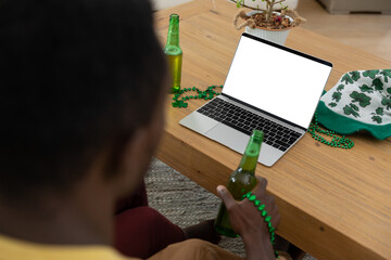 African american man with beer on st patrick's day video call using laptop with copy space on screen