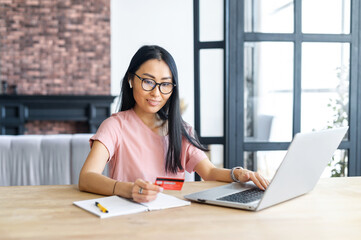 Nice young Asian woman is paying with debit card online on the laptop, a smiling girl is shopping online, making purchase, using app on the computer for e-banking sitting at the desk in home office