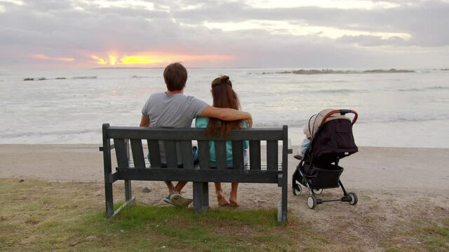 Rear view of loving family couple sitiing by a ocean looking at sunset with baby stroller.