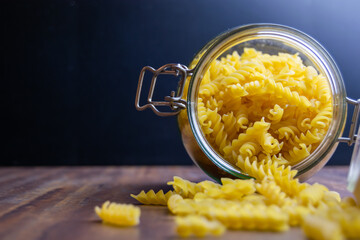 Fussili pasta falling out from a big air tight glass jar. Raw dry pasta in zero waste glass container. Messy food composition on dark background with selective focus and copy space.