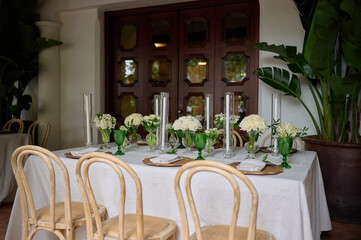 Table setting with serviette and greens, wooden plates and green glasses, with vases with white flowers and candles in the room