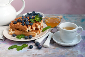 Belgian waffles with honey, blueberries, bananas, and mint on a light grey background. Breakfast with a croissant and a cup of green tea with lemon