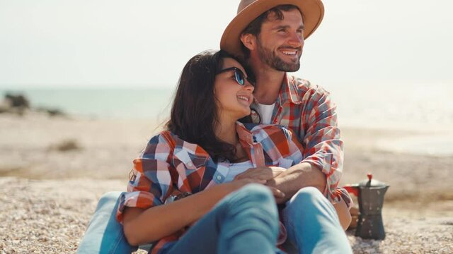 happy romantic couple sits on sea beach, talks, laughs and hugs. handsome adult man in casual checkered shirt and hat embracing beautiful charming young woman. moments of love, tender and happiness