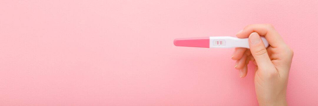 Young adult woman hand holding pregnancy test with two stripes on light pink table background. Pastel color. Positive result. Closeup. Wide banner. Empty place for text. Copy space. Top down view.