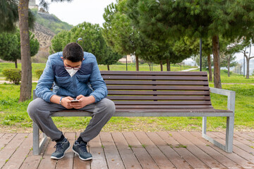 Fototapeta premium Young boy wearing face mask due to coronavirus pandemic sitting in bench and using his mobile phone.