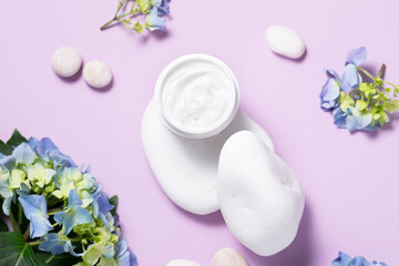 Obraz na płótnie Canvas Creative composition with organic moisturizing cream on the podium made with decorative white stones and fresh hydrangea flowers on light lilac background top view. Skin care products.