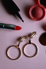 Gold vintage earrings, red lipstick, eyeliner and blush on pink background. Top view.