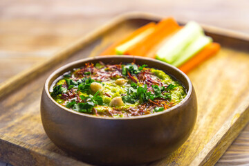 Homemade chickpea green hummus with herbs in round bamboo bowl, celery, carrot sticks on wood