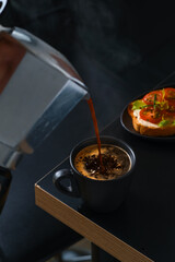 Hot fresh espresso coffee is poured into a cup from a geyser coffee maker. Bar table, cafe breakfast idea, cheese and tomato toast or antipasto. Dark moody background