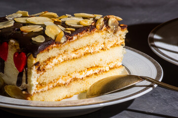 Piece of layered homemade sponge cake on a white saucer with a spoon and a linen napkin, black background, hard light, photo in a low key.