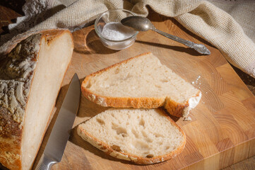 Still life - yeast-free buckwheat bread with sliced pieces, and coarse salt in a glass jar, a knife, and a linen napkin on a wooden board, a wooden background, hard light, a photo in a low key.