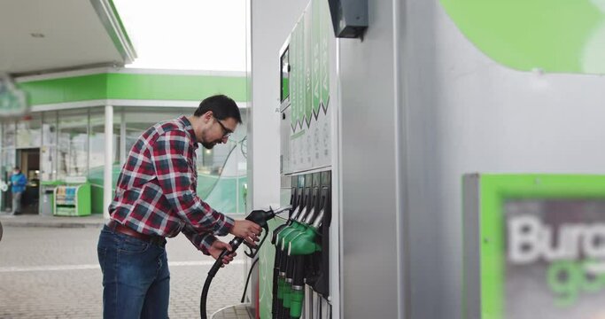 Portrait of a Young Man in Glasses, Stylishly Dressed, Holding a Car Refueling Gun. Shooting At the Gas Station. Fuel nozzle getting put into its place. Gasoline, gas, fuel, petroleum concept.