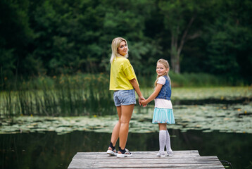 Young family, mother and daughter stand on a wooden pier holding hands, turning around and looking...