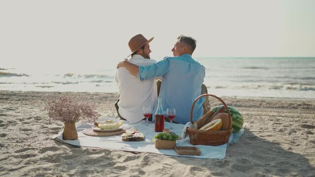 Homosexual lgbt couple, gay men having picnic at the beach, sitting on blanket and embracing, looking to ech other with love. Lgbt life