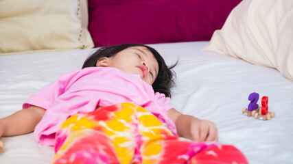 Little Asian cute girl resting on bed in bedroom with her wooden number train toy on side. Child fell asleep while lying on the bed. Kids aged 4 years old.