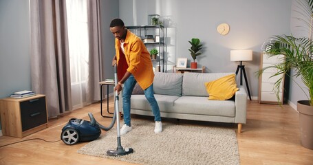 African American busy handsome young guy vacuuming cozy living room doing housework, male cleaning house using vacuum keeping home clean, housekeeper, everyday life concept - obrazy, fototapety, plakaty