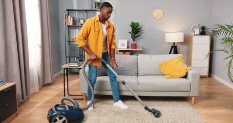 Handsome African American young concentrated busy man vacuuming cleaning modern living room, male...
