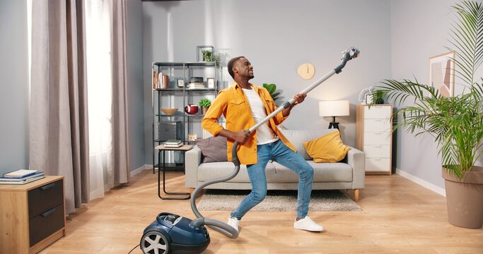 African American joyful happy handsome young guy vacuuming living room at home doing housework dancing having fun pretending to play on guitar, male cleaning house using vacuum, housework concept