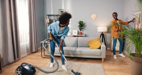 African American young nice happy married couple cleaning living room in apartment, wife vacuuming carpet floor while husband wiping dust on furniture behind, family concept, routine life