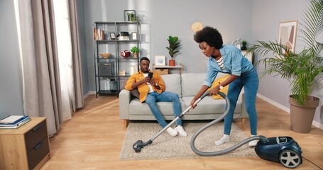 Fototapeta na wymiar Happy beautiful young African American woman vacuuming carpet while handsome beloved man resting on couch texting on smartphone surfing online social network app, cleaning home concept