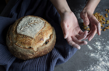 Top view photo of a table. Round sourdough bread  made of rye grain, navy blue kitchen towel, woman's hands holding bread. Homemade bread recipes. 
