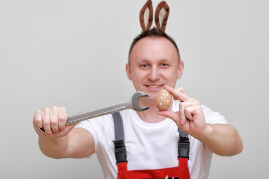 Easter holiday, celebration concept. funny smiling engineer worker or mechanic is wearing rabbit ears on head, holding wrenches and painted egg white background. celebrating orthodox day