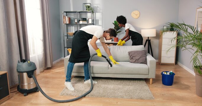Busy young mixed-races male and female professionals working in aprons cleaning apartment, male cleaner vacuuming sofa while woman mopping floor in client's home, cleaning industry concept