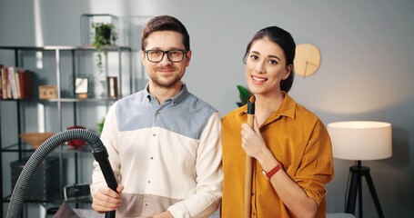 Portrait of cheerful Caucasian happy young married couple wife and husband dancing and singing while cleaning apartment with vacuum cleaner and mop smiling to camera, family having fun together