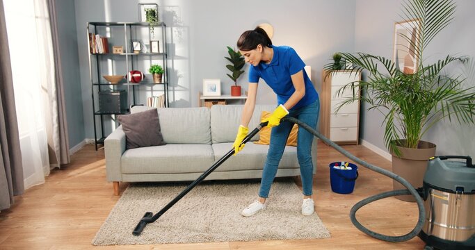 Beautiful Caucasian young woman in yellow gloves vacuuming carpet floor at home in living room. Pretty female uses vacuum cleaner in apartment, domestic work, housekeeper concept