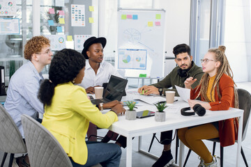 Group of five multiracial young people in casual outfit sitting at modern office and discussing new startup project. Creative students sharing with business ideas during meeting.