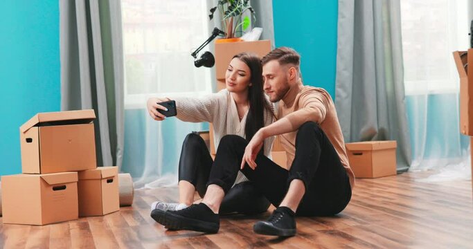 Happy young family with cardboard boxes moving in new home, sitting together on the floor in living room, taking selfie picture, hugging and smiling wide.