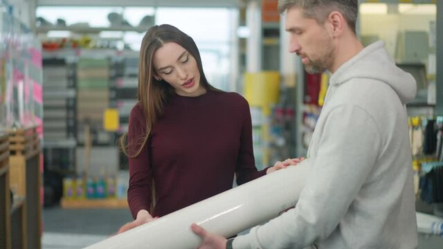 Caucasian man walking with roll of plastic construction film to woman in hardware store. Husband choosing supplies for repair or moving with wife in shop indoors. Buying concept.