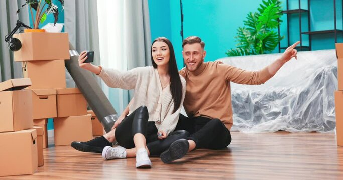 Young couple move into new apartment. Camera moves fowards from people sitting on floor and taking selfie. Making funny facial expressions and posing. Happy people after relocation.
