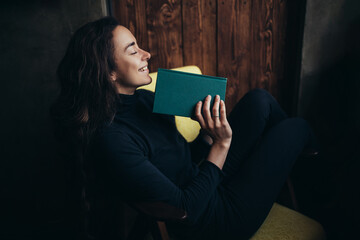Young beautiful woman with long curly hair sitting in a comfortable armchair and reading interesting book.