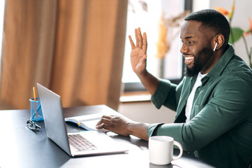 Online communication. Happy smiling African American guy, freelancer or student, waving hand, greeting, uses laptop for video conference with colleagues - obrazy, fototapety, plakaty