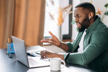 Displeased young African American man, displeased looks at the laptop, disagreeing with colleague during video call, sitting at his desk, gesturing hand