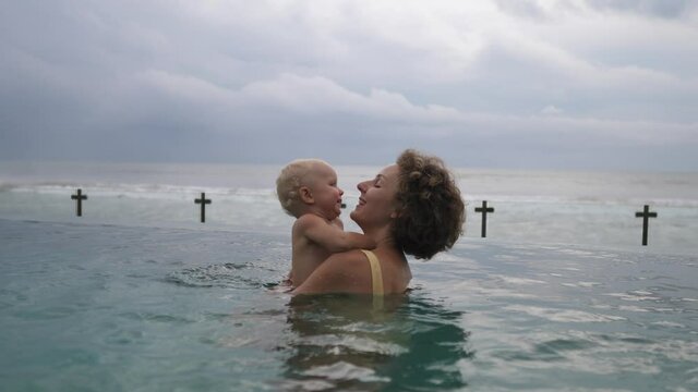 Young Caucasian mother plays with her baby in the swimming pool overlooking an ocean. Happy childhood concept