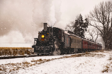 Fototapeta na wymiar View of An Antique Restored Steam Locomotive Blowing Smoke and Steam Traveling Thru Farmlands and Countryside in a Snow Storm