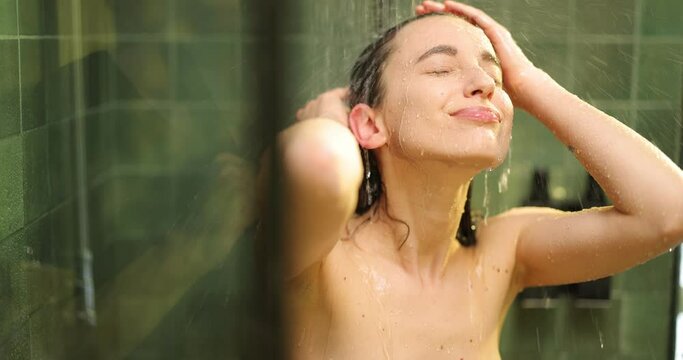 Young adult woman taking a rain shower, enjoying water splashes on her face and hair in the green bathroom