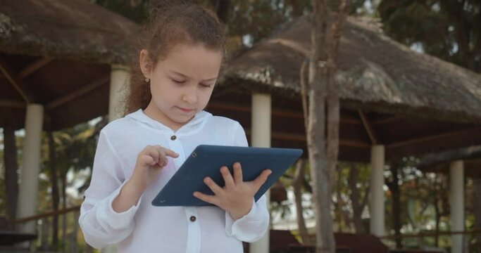 Curious cute girl 7-8 years old is having fun with a digital tablet device watching a cartoon while walking on the beach. The child has a rest plays in the tablet outdoors.