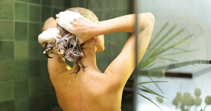 Young woman takes a shower, soaping her brown hair with a shampoo in a green bathroom. View from the backside
