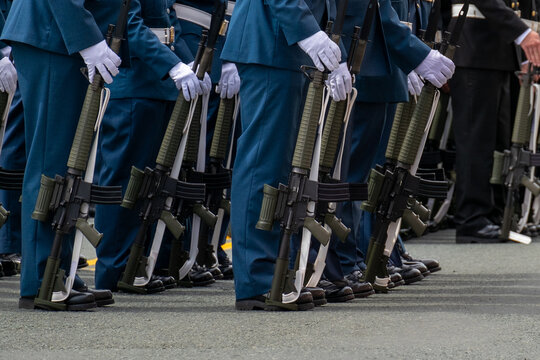 Soldiers standing at attention with guns or rifles at their feet. The men are in blue uniforms wearing white gloves. They are standing at a parade in dress uniform. The view is from the waist down.
